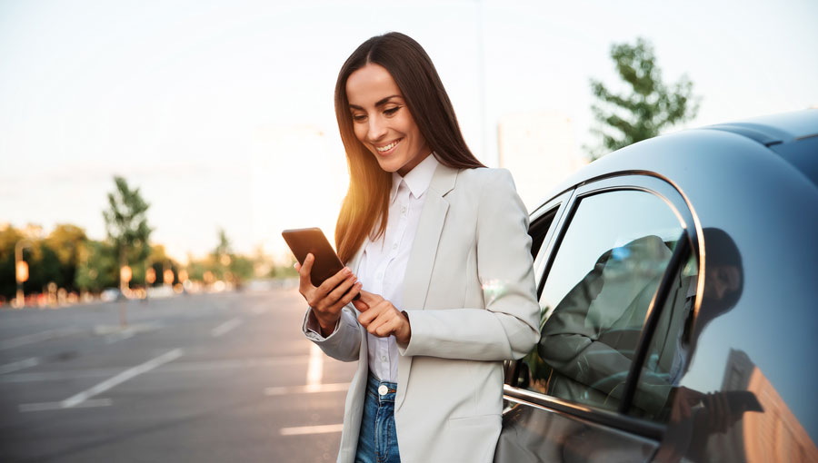 Smiling woman looking at phone app from VFCU Harlingen credit union near modern car outdoors.